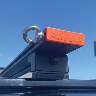 An orange Alu-Cab roof rack load bar plastic end cap with a silver metal eye bolt, mounted on a black roof rack bar against a blue sky.